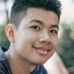 Cheerful Asian teenage boy in dark shirt with short black hair looking at camera with friendly toothy smile in forest on blurred background